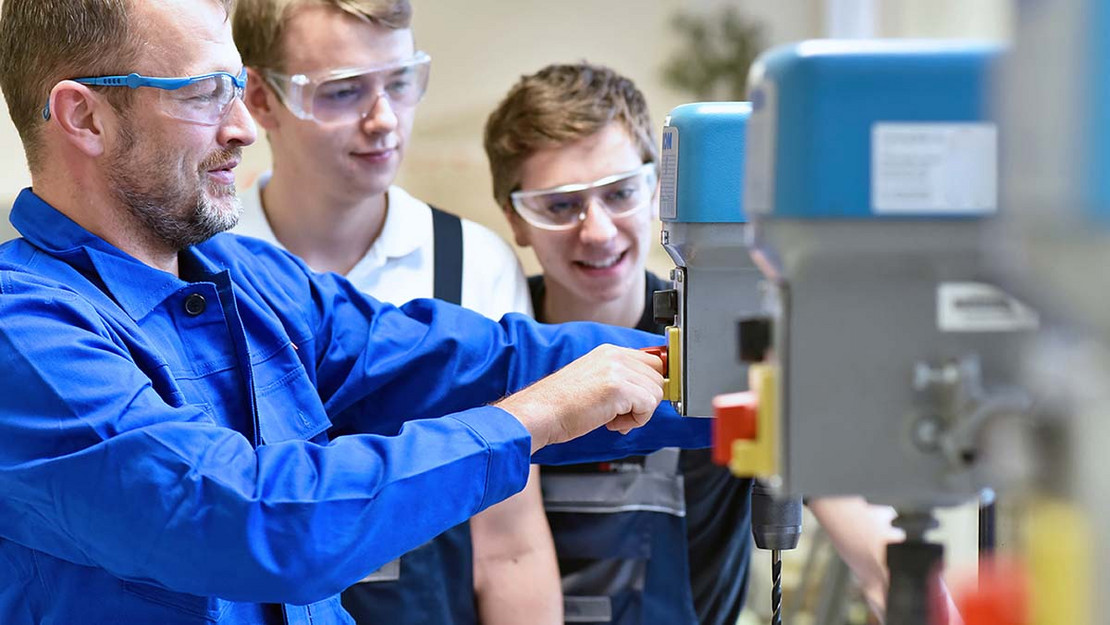 Ein älterer und zwei jüngere Männer stehen in einer Werkhalle an Maschinen. 