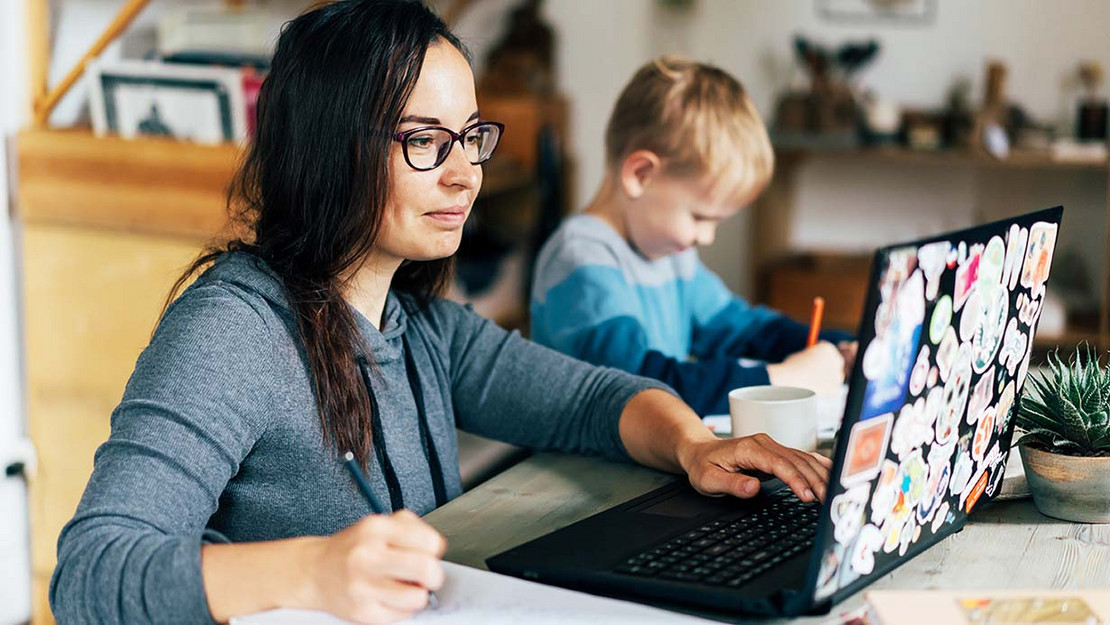 Eine Frau arbeitet zu Hause an einem Laptop während ihr Kind daneben sitzt. 