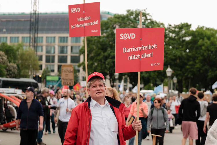 Mann hält ein Schild mit der Aufschrift "Barrierefreiheit nützt allen" in die Höhe. 