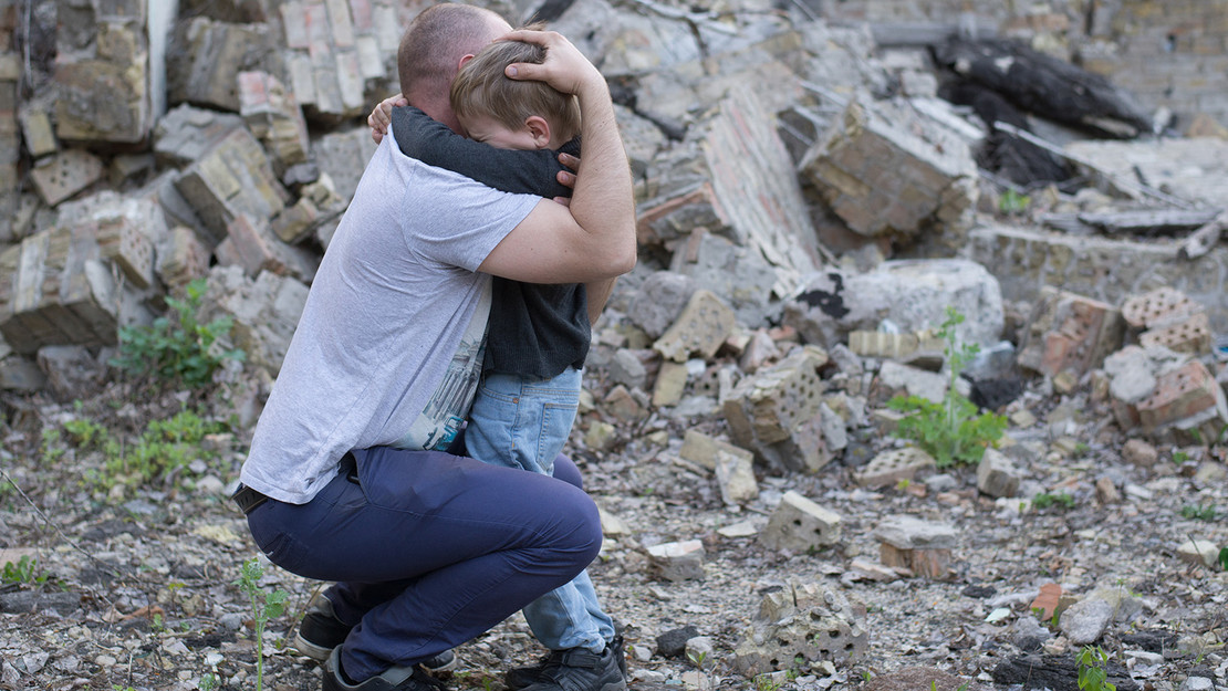 Vater umarmt sein kleines Kind vore Trümmern. 