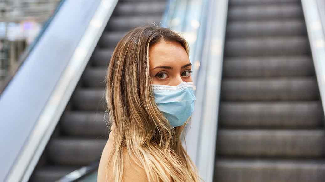 Frau mit Maske vor einer Rolltreppe. 